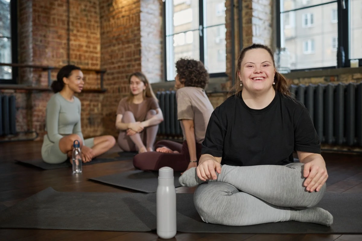 Disabled woman with down syndrome in yoga class with legs crossed smiling at camera with friends in the background